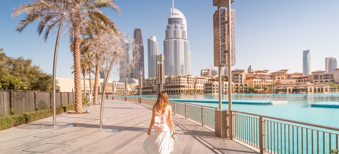 Eine Frau in weißem Kleid spaziert an einer sonnigen Uferpromenade in Dubai entlang. Im Hintergrund ragen moderne Wolkenkratzer in den blauen Himmel, Palmen säumen den Weg, und das Wasser einer künstlichen Lagune glänzt türkis im Sonnenlicht.