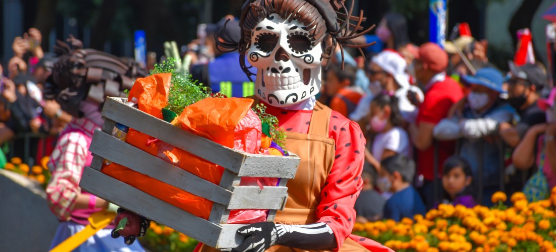 Eine festlich gekleidete Teilnehmerin der Día de los Muertos Parade trägt eine kunstvolle Calavera-Maske und ein traditionelles Kleid in lebendigen Farben. Sie hält eine mit Blumen und bunten Stoffen dekorierte Kiste, während im Hintergrund eine jubelnde 