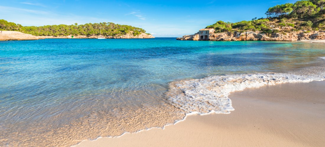 Eine idyllische Bucht mit feinem, hellem Sandstrand und türkisblauem Wasser. Die Bucht ist von sanft ansteigenden Felsen und grünen Pinienbäumen umgeben. Auf der linken Seite erstreckt sich eine kleine felsige Landzunge ins Meer. Rechts im Hintergrund bef