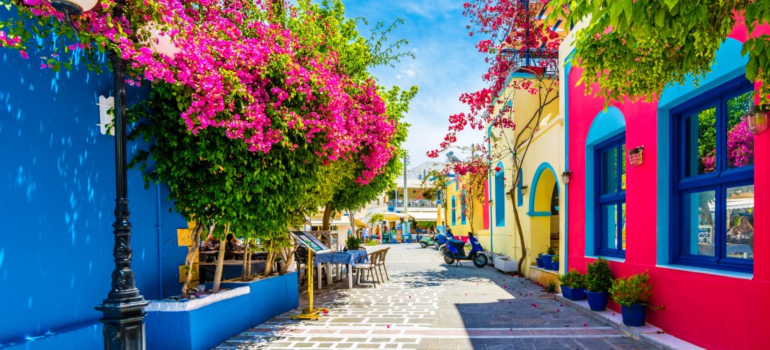 Lebendige Gasse in einer mediterranen Stadt mit bunten Häuserfassaden in Blau, Gelb und Pink. Über der Straße hängen üppige Bougainvillea mit leuchtend pinken Blüten. Auf der linken Seite stehen Tische und Stühle eines kleinen Straßencafés, im Hintergrund
