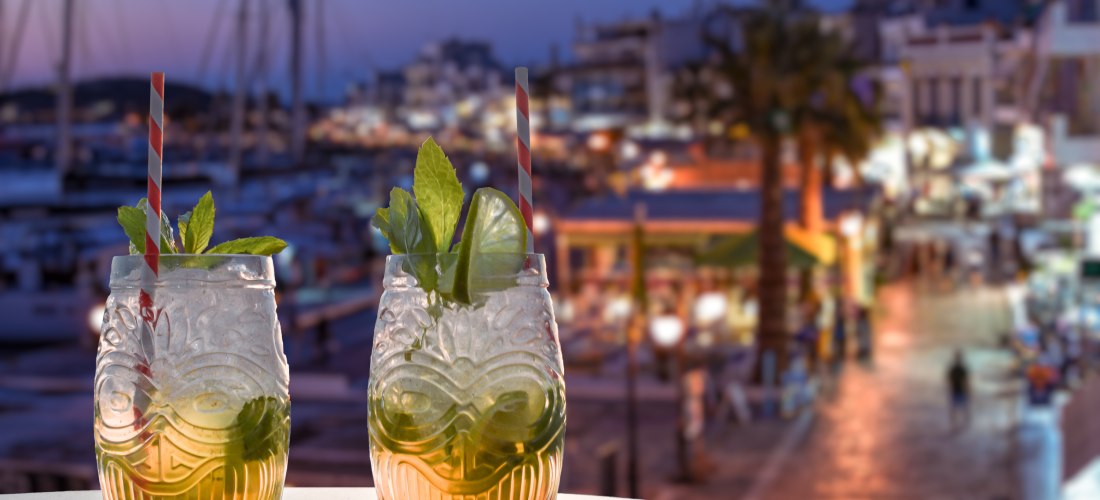 Zwei Cocktails in verzierten Gläsern mit Minzblättern, Limettenscheiben und gestreiften Trinkhalmen stehen auf einem Tisch. Im Hintergrund unscharf eine abendliche Hafenpromenade mit Lichtern, Restaurants und Bootsmasten.
