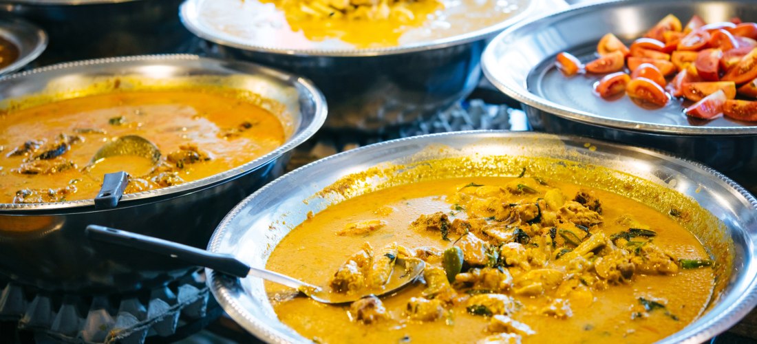 Das Bild zeigt mehrere große silberne Metallschalen, gefüllt mit typischen thailändischen Currys und Zutaten. Im Vordergrund befinden sich zwei gelb-orangene Currys mit Gemüse, Kräutern und Fleischstücken, aus denen große Löffel ragen. In weiteren Schale