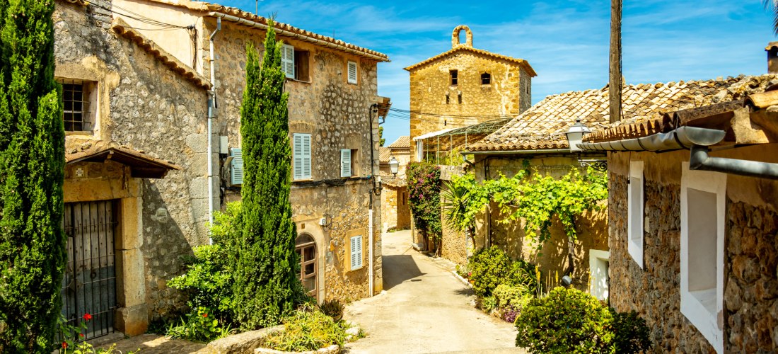 Ein malerisches Dorf auf Mallorca mit traditionellen Steinhäusern, grün gestrichenen Fensterläden und engen Kopfsteinpflastergassen. Die Häuser sind von grünen Pflanzen und bunten Blumen geschmückt. Eine kleine Kirche mit einem Glockenturm erhebt sich im 