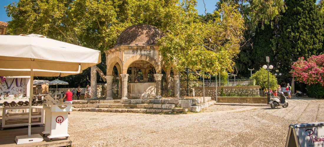 Platz mit einem überdachten achteckigen Brunnen unter einer alten Platane, umgeben von Steinmauern und Säulen. Im Vordergrund ein Verkaufsstand unter einem Sonnenschirm, im Hintergrund große Bäume und Menschen auf dem Platz. Der Ort ist als Platanenplatz 