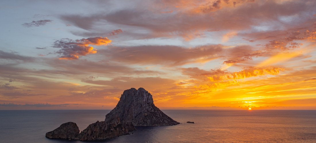 Menschen beobachten den Sonnenuntergang mit Blick auf die Felseninsel Es Vedrà. Der Himmel ist orange-rot gefärbt, die Sonne versinkt im Meer hinter der markanten Insel.