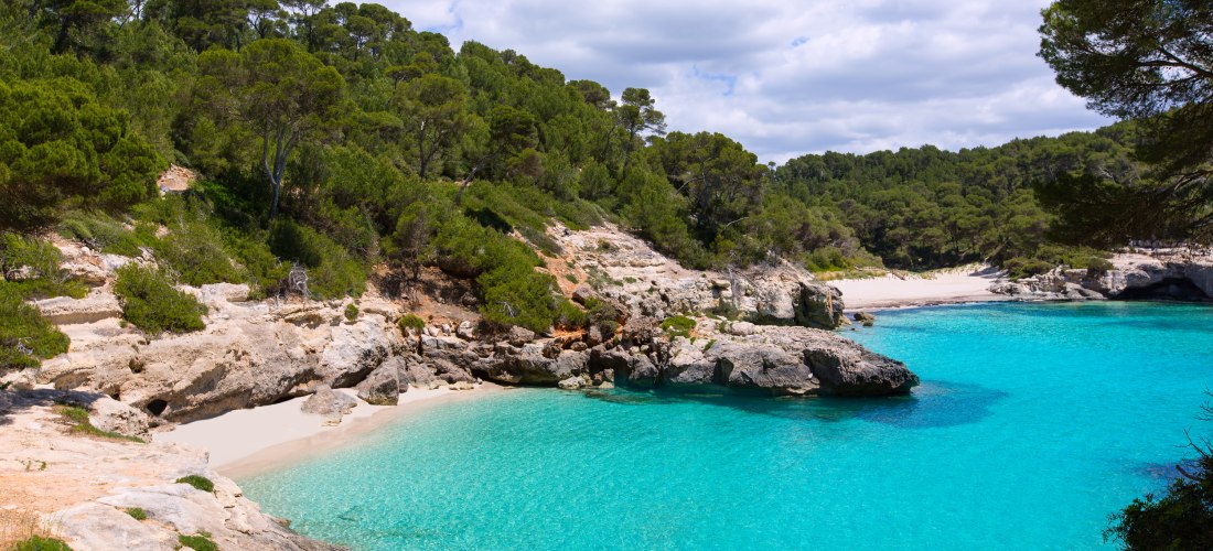 Kleine Badebucht Cala Mitjana und Mitjaneta auf Menorca: klares, türkisfarbenes Wasser, helle Felsen und dichter Pinienwald, die den weißen Sandstrand umrahmen.
