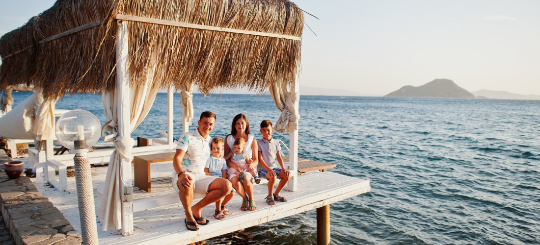 Eine Familie mit fünf Personen sitzt unter einer strohgedeckten Strandhütte auf einem Holzsteg direkt am Meer. Im Hintergrund ist eine kleine Insel zu sehen, die Sonne scheint und die Stimmung ist entspannt und sommerlich.