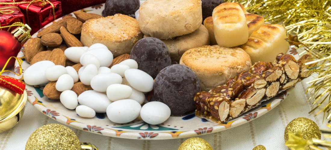 Ein dekorativer Weihnachtsteller mit typisch spanischem Weihnachtsgebäck: Polvorones, Turrón, Marzipanfiguren, kandierten Mandeln und Schokoladenkugeln, umrahmt von goldenen Christbaumkugeln und Weihnachtsdekoration.