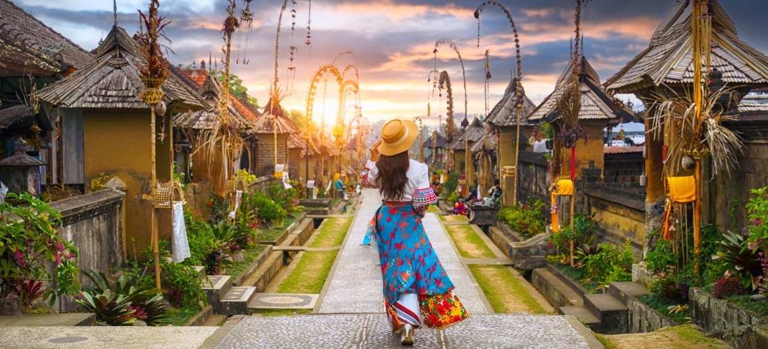 Eine Frau mit Hut und farbenfrohem, traditionell inspiriertem Kleid spaziert eine gepflasterte Straße entlang durch das Dorf Penglipuran auf Bali. Links und rechts stehen kleine balinesische Häuser mit kunstvoll geschmückten Toren und blühenden Gärten. Ho
