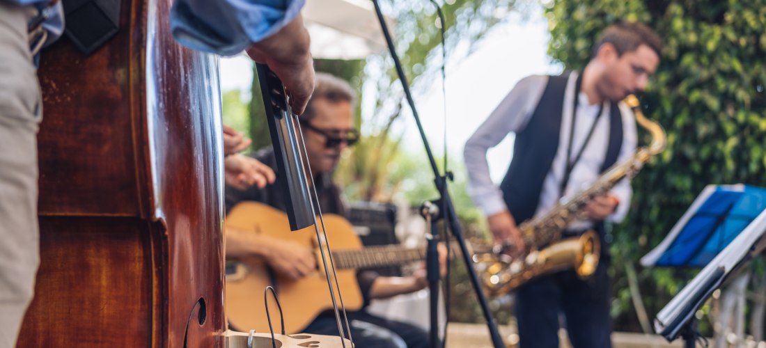 Eine Jazzband spielt im Freien. Im Vordergrund ein Kontrabass, im Hintergrund ein Gitarrist und ein Saxophonist. Die Musiker sind entspannt und konzentriert