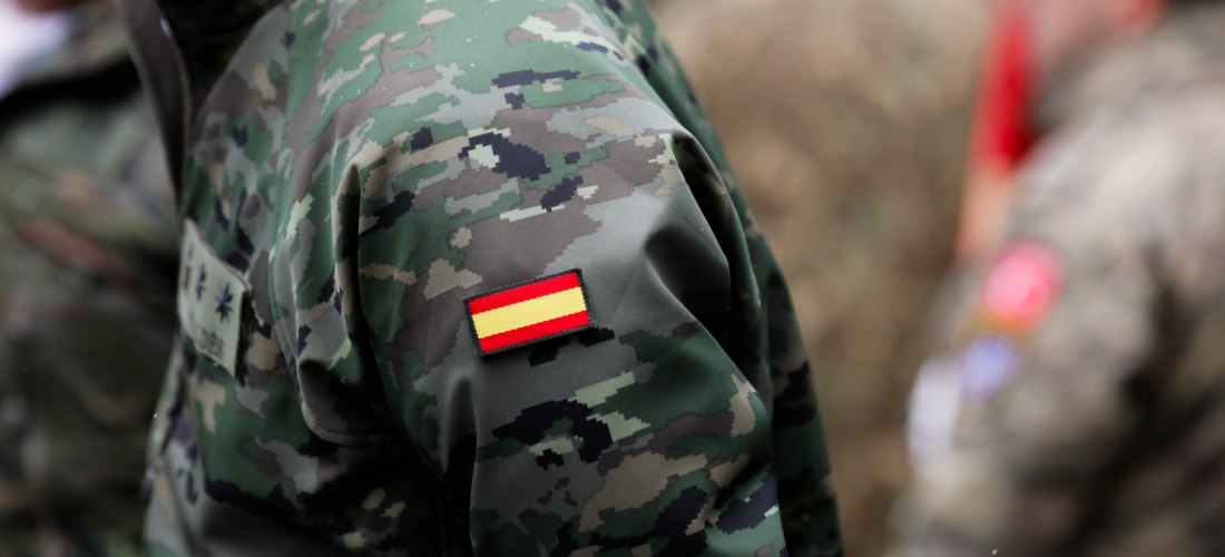 Nahaufnahme einer militärischen Tarnuniform mit einem Spanien-Patch am Oberarm. Im Hintergrund sind unscharf weitere Soldaten in ähnlicher Kleidung zu sehen.