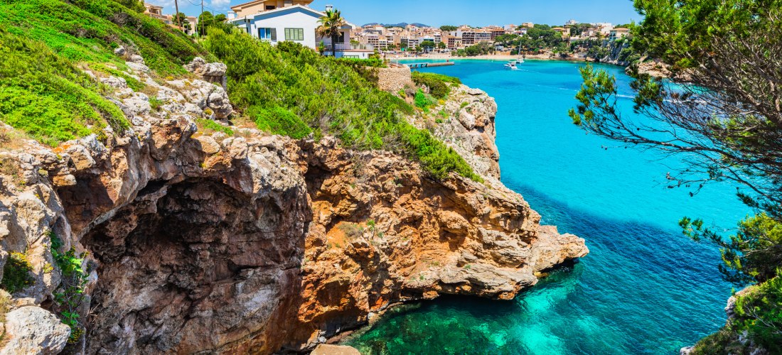 Panoramaaufnahme von Porto Cristo mit dem türkisfarbenen Hafenbecken und der Stadt dahinter. Weiße und sandfarbene Häuser reihen sich dicht aneinander, viele mit Flachdächern oder Dachterrassen. Im Hafen liegen zahlreiche Boote und Yachten. Rechts führt e
