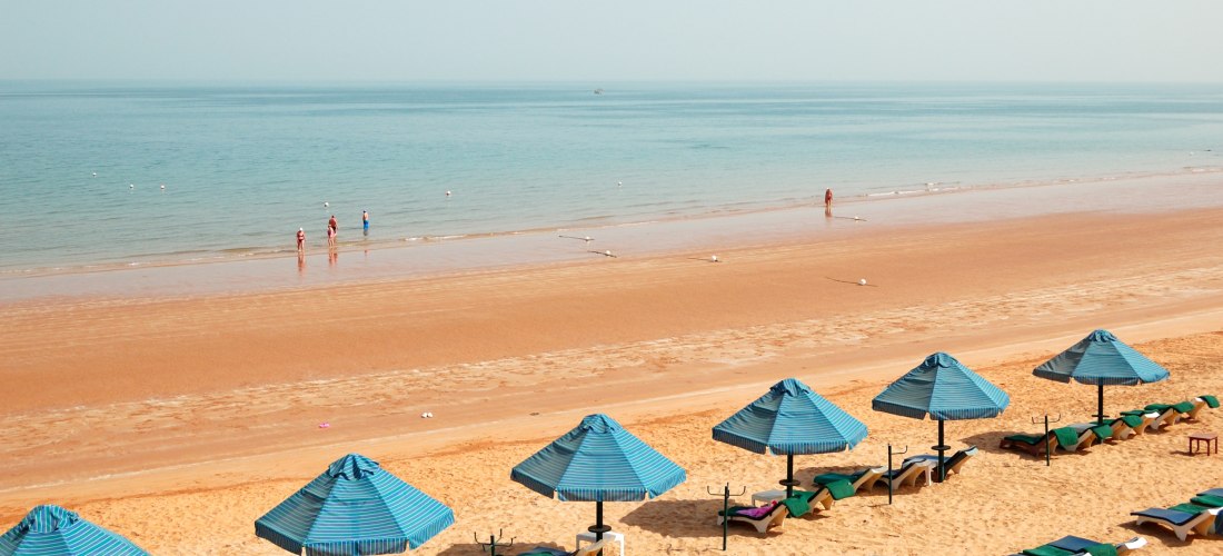 Ruhiger Sandstrand mit blauen Sonnenschirmen und Liegen, Menschen spazieren am flachen Ufer