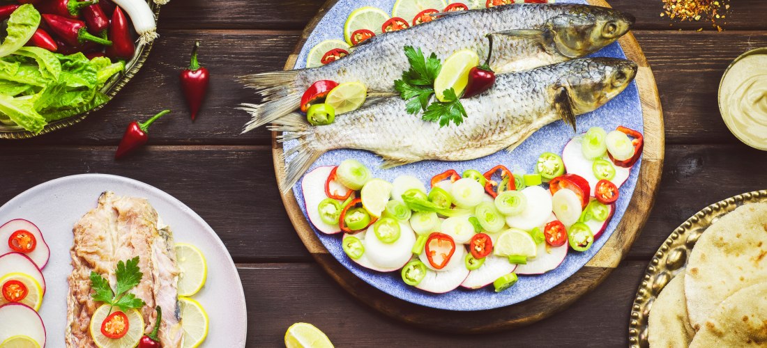 Zwei ganze gesalzene Fische auf einem blauen Teller, umgeben von Limettenscheiben, Chili, Frühlingszwiebeln und Radieschen. Daneben verschiedene Beilagen wie Fladenbrot, Zitronenschnitze und Gewürze, auf einem dunklen Holztisch angerichtet.