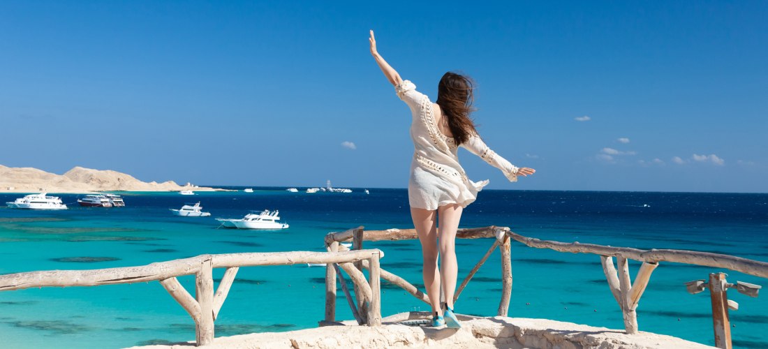 Eine Frau steht auf einer kleinen Sandterrasse oberhalb des Meeres und blickt in Richtung Wasser. Sie trägt ein weißes, luftiges Strandkleid, ihre Haare wehen im Wind. Vor ihr liegt das azurblaue Meer, gesäumt von mehreren Yachten und Booten, im Hintergru