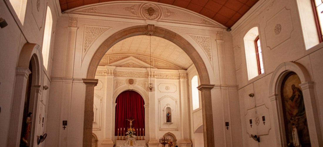 Innenansicht einer katholischen Kirche mit hohen weißen Wänden, großen Rundbogen, klassizistischen Säulen und einem roten Vorhang hinter dem Altar. Der Altarraum ist geschmückt mit Kerzen und Blumen, einige Menschen sitzen in den Bänken