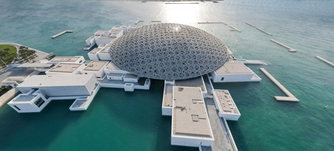 Luftaufnahme des Louvre Abu Dhabi mit seiner ikonischen Kuppel, die auf dem Wasser zu schweben scheint, mit Blick auf die Skyline von Abu Dhabi im Hintergrund