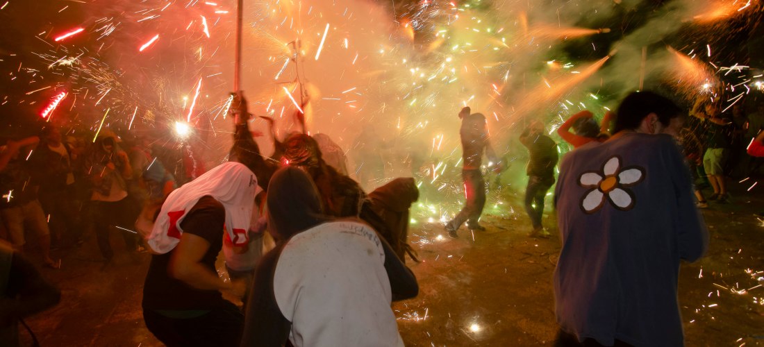 Menschen tanzen und ducken sich inmitten eines spektakulären Funkenregens während eines Correfoc, eines traditionellen Feuerlaufs auf Mallorca. Pyrotechnische Effekte sprühen in alle Richtungen, es ist Nacht. Einige tragen Kapuzen oder haben sich mit Tüch
