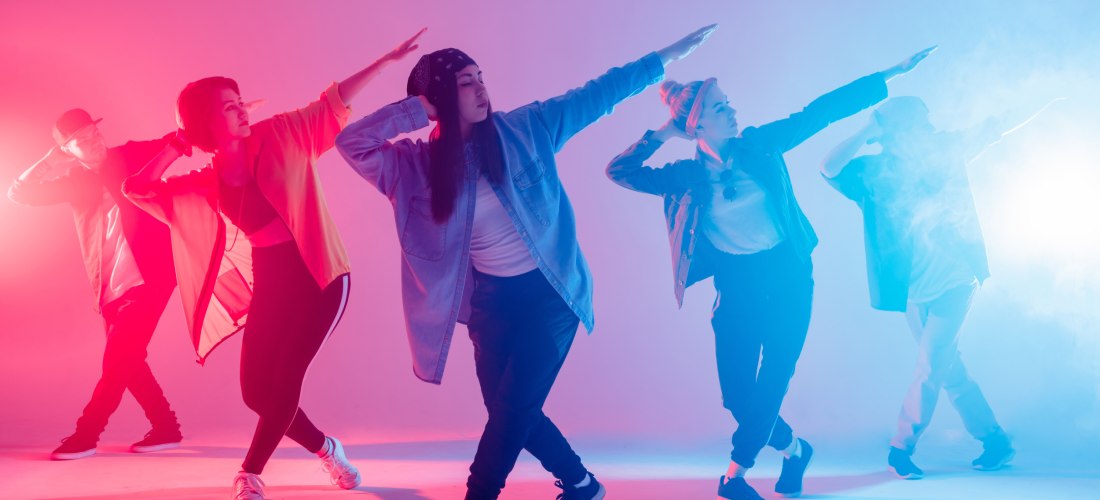 Fünf junge Tänzer:innen in Streetwear posieren synchron unter buntem pink-blauen Bühnenlicht – energiegeladene Hip-Hop-Atmosphäre im Studio