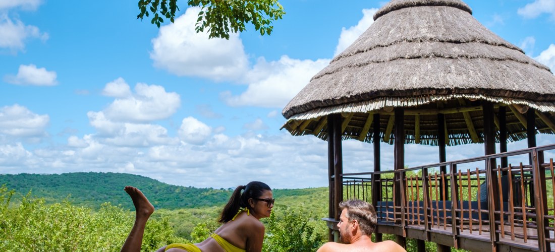 Ein Paar entspannt gemeinsam in einem Infinity-Pool mit Blick ins grüne Buschland. Die Frau liegt auf dem Bauch, der Mann lehnt am Beckenrand. Im Hintergrund steht eine Hütte mit Strohdach, darüber ein blauer Himmel mit weißen Wolken.