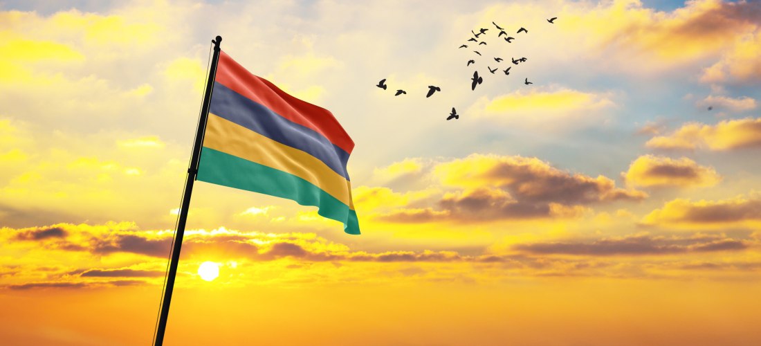 Dieses Bild zeigt die Flagge von Mauritius, die stolz im Wind weht, während die Sonne am Horizont untergeht. Der Himmel ist in warmen Gelb-, Orange- und Blautönen gefärbt, mit verstreuten, weichen Wolken, die von der Abendsonne beleuchtet werden. In der r