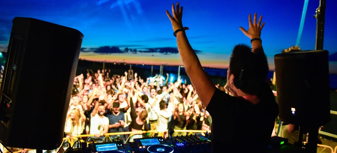 DJ steht auf einer Bühne mit erhobenen Händen vor einer großen feiernden Menschenmenge. Im Vordergrund Mischpulte und Lautsprecher. Der Himmel im Hintergrund zeigt ein spektakuläres Farbspiel zwischen Blau und Orange beim Sonnenuntergang.