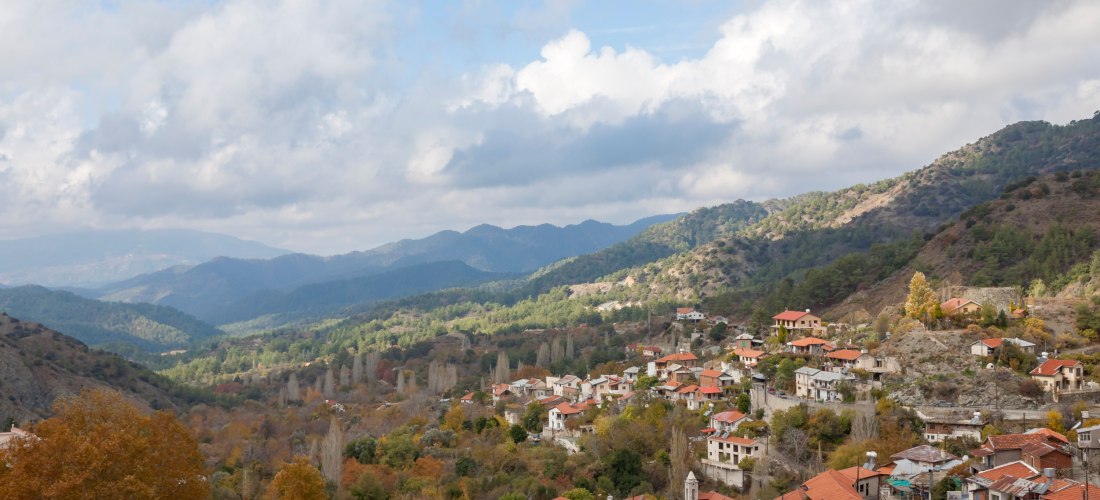 Panoramablick auf ein traditionelles Bergdorf im Troodos-Gebirge mit roten Ziegeldächern, bewaldeten Hängen und Wolken über den Bergen.