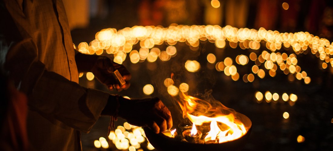 das eindrucksvolle Bild zeigt eine spirituelle Zeremonie, bei der ein Teilnehmer kleine Öllampen (Diyas) in einer großen Metallschale entzündet. Im Hintergrund leuchten zahlreiche weitere Kerzen und Lampen in einem warmen, goldenen Schein und erzeugen ein