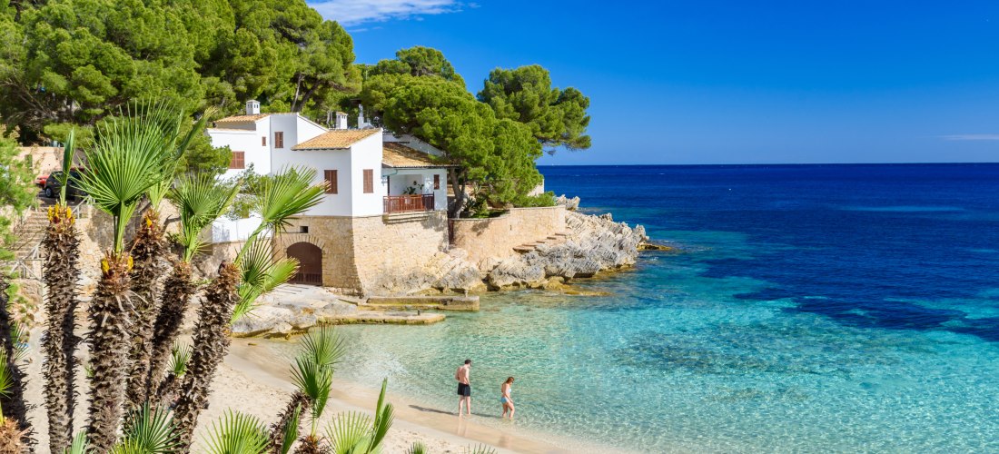 Idyllische Bucht auf Mallorca mit kristallklarem, türkisblauem Wasser. Am feinsandigen Strand spazieren zwei Personen barfuß am Wasser entlang. Im Hintergrund stehen weiße Häuser mit Terrakotta-Dächern und braunen Fensterläden auf einem Felsvorsprung, umg