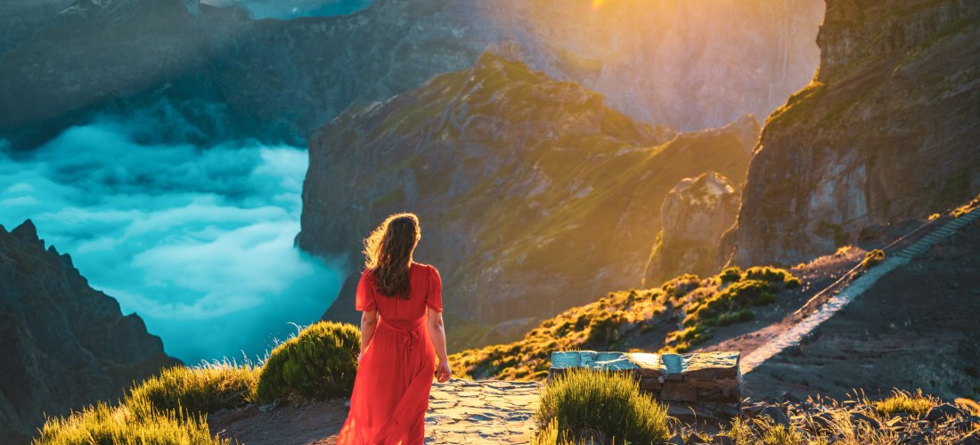 Frau im roten Kleid steht barfuß auf einem Bergpfad auf Madeira und blickt in ein dramatisches Tal, in dem sich Wolken unter ihr sammeln. Die Szene ist in goldenes Morgenlicht getaucht, umgeben von steilen Felswänden und grünen Berghängen