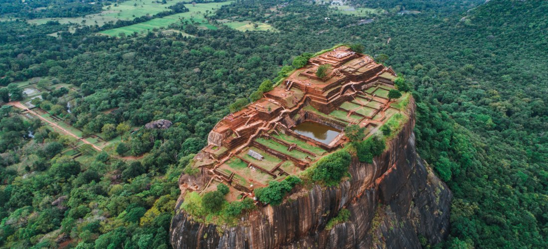 Luftaufnahme der Felsenfestung Sigiriya in Sri Lanka. Der gewaltige Felsen ragt aus dem dichten Dschungel heraus und zeigt Reste einer alten Palastanlage mit Mauern, Wasserbecken und Terrassen. Umgeben von grünem Wald und offenen Landschaften.
