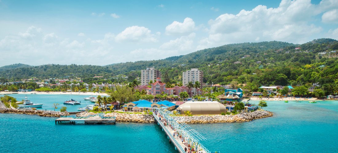Blick auf die tropische Küstenstadt Ocho Rios in Jamaika. Im Vordergrund führt ein langer Steg über das türkisfarbene Meer zur Anlegestelle, wo Menschen entlanglaufen. Am Ufer befinden sich bunte Gebäude, Palmen und kleine Yachten in der Bucht. Dahinter e