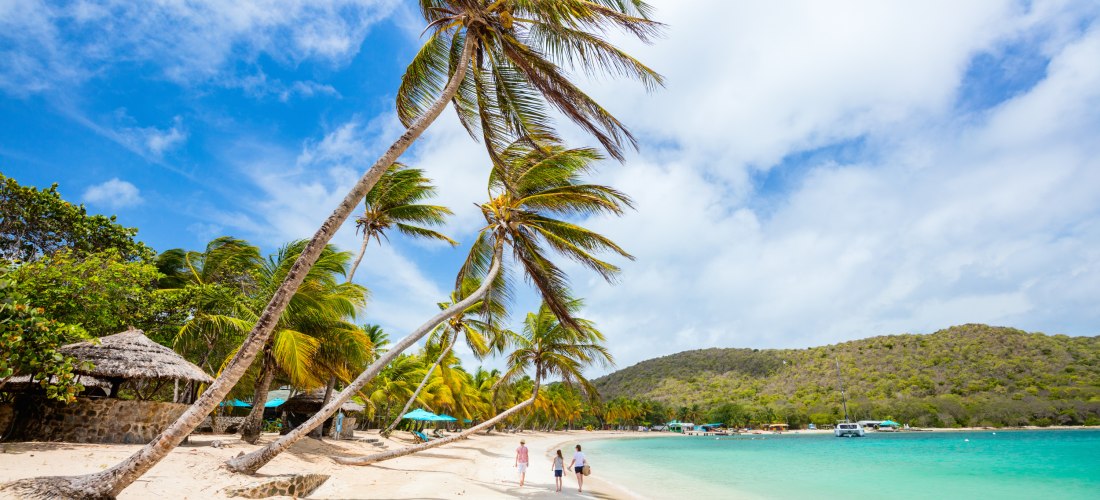 Breiter Sandstrand mit sanft geschwungenen Palmen, die sich in den blauen Himmel neigen. Drei Menschen spazieren entspannt am türkisfarbenen Meer entlang. Im Hintergrund liegen grüne Hügel und einige Boote vor Anker. Der Himmel ist leicht bewölkt und stra