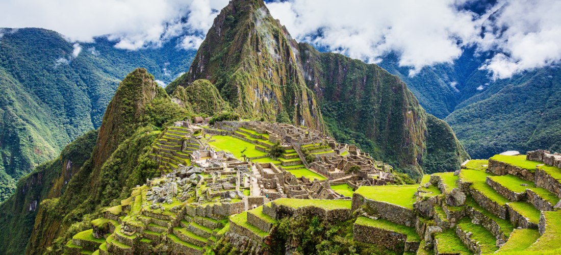 Panoramablick auf die Ruinen von Machu Picchu in Peru, umgeben von üppig grünen Andenbergen. Die steinernen Terrassen und Bauwerke der Inka-Stadt liegen auf einem Bergrücken, im Hintergrund ragt ein spitzer, bewaldeter Berg auf. Der Himmel ist leicht bewö