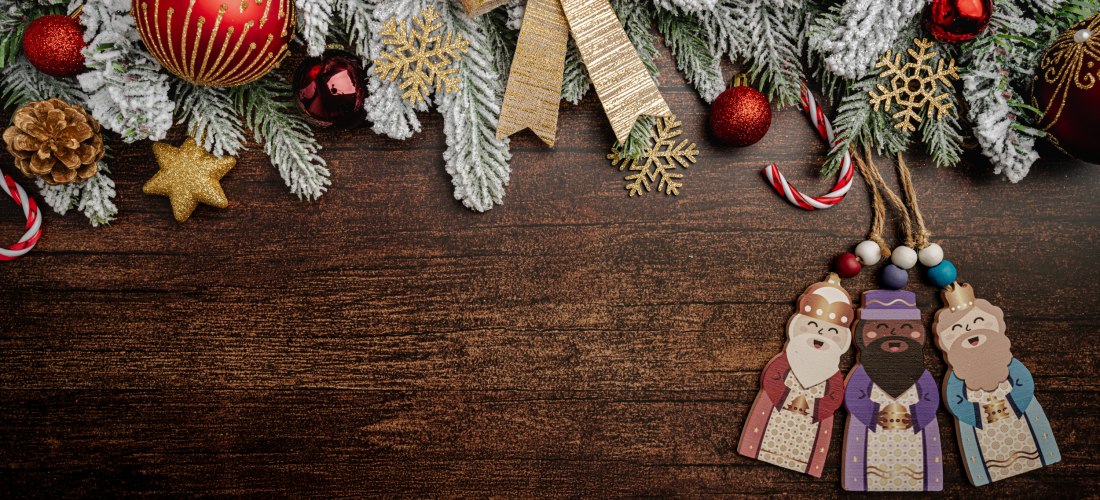 Oben im Bild hängt ein weihnachtlicher Kranz mit verschneiten Tannenzweigen, roten Kugeln, Beeren, goldenen Schneeflocken und einer großen goldenen Schleife. Unten rechts liegen drei weihnachtliche Holzfiguren der Heiligen Drei Könige auf einer dunklen Ho