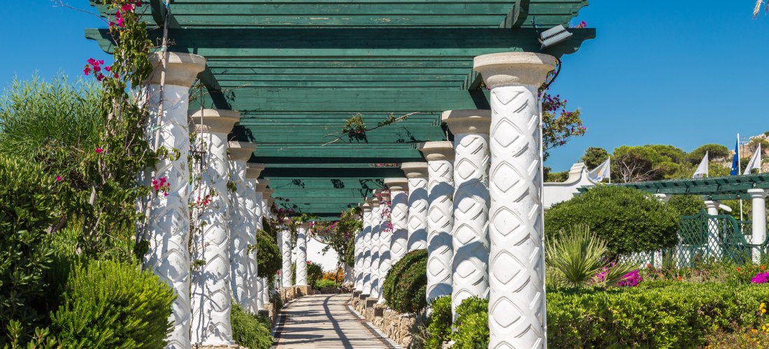 Gepflasterter Weg unter einer überdachten Pergola mit weißen, kunstvoll verzierten Säulen und grünem Holzdach. Kletterpflanzen mit pinkfarbenen Blüten umranken die Struktur. Links und rechts des Weges befinden sich dicht bepflanzte mediterrane Gärten mit 