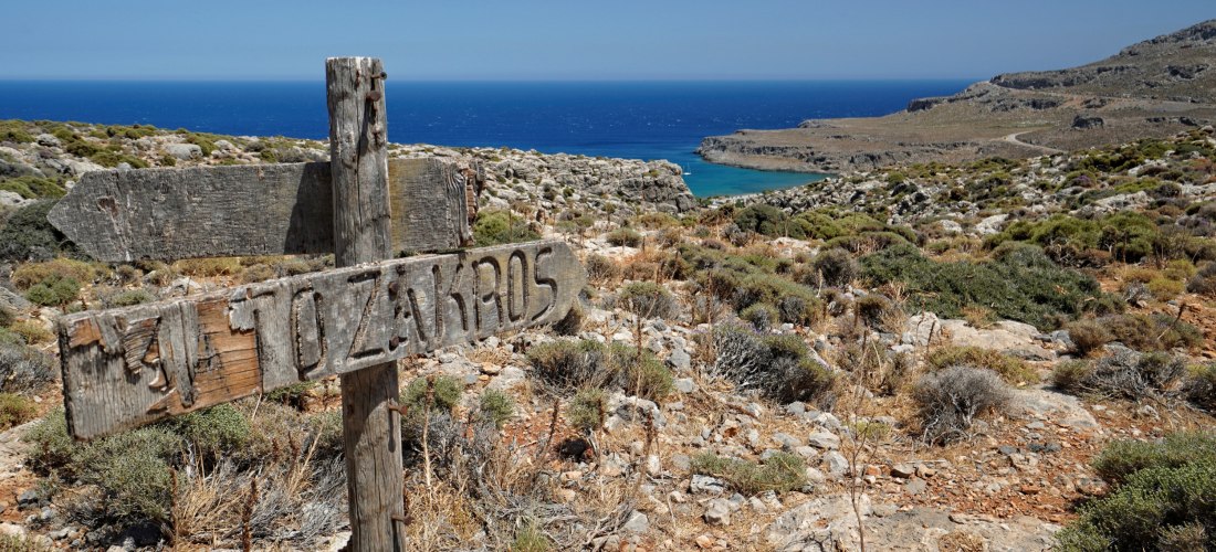 Verwittertes Holzschild mit der Aufschrift 'Zakros' in einer felsigen Landschaft mit Blick auf das blaue Meer auf Kreta