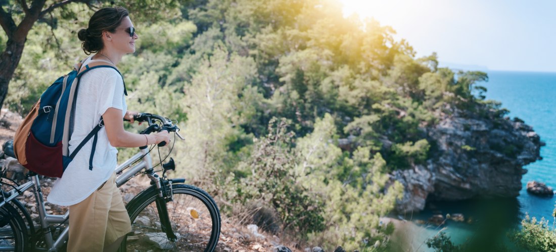 Eine Frau mit Rucksack steht mit ihrem Fahrrad an einem Aussichtspunkt im Wald. Sie blickt lächelnd auf das Meer und eine bewaldete Felsenküste. Die Sonne scheint durch die Bäume, es herrscht eine entspannte, sommerliche Atmosphäre.
