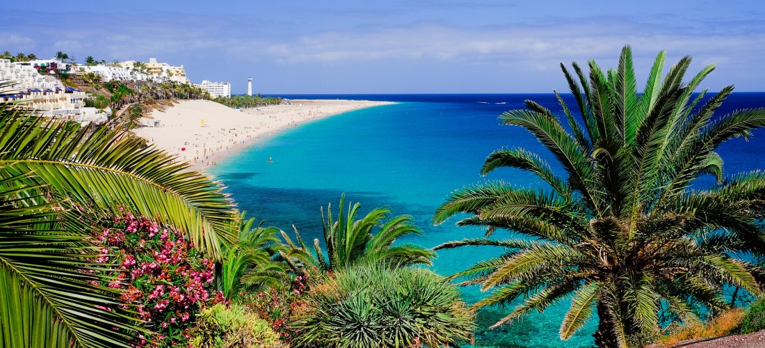 Blick auf einen langen, hellen Sandstrand mit türkisblauem Meer, gesäumt von Palmen, tropischen Pflanzen und Hotelanlagen. Im Hintergrund ein Leuchtturm am Horizont.