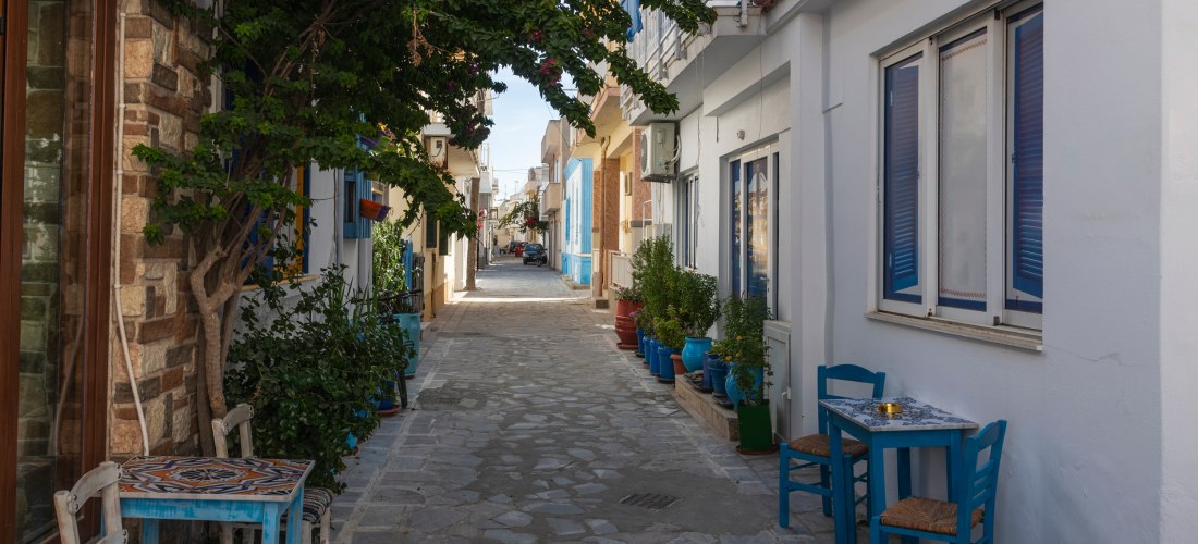 Schmale Gasse auf Kos mit traditionellen weißen Häusern, bunten Fensterläden, Blumen und kleinen Cafétischen mit blauen Stühlen