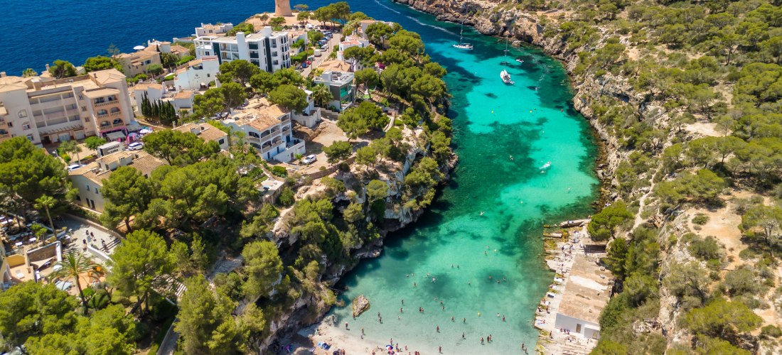 Eine atemberaubende Bucht mit türkisblauem Wasser auf Mallorca, umgeben von steilen Felsklippen mit üppigen Pinienwäldern. Kleine Boote liegen vor Anker, während Menschen an einem hellen Sandstrand baden und sonnen. Im Hintergrund sieht man eine Siedlung 
