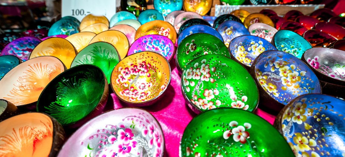 Das Bild zeigt eine große Auswahl handbemalter Kokosnussschalen, die auf einem Marktstand ausgestellt sind. Die Schalen sind leuchtend bunt in Farben wie Grün, Pink, Blau, Gelb und Lila und mit floralen oder abstrakten Mustern verziert. Sie glänzen im Lic