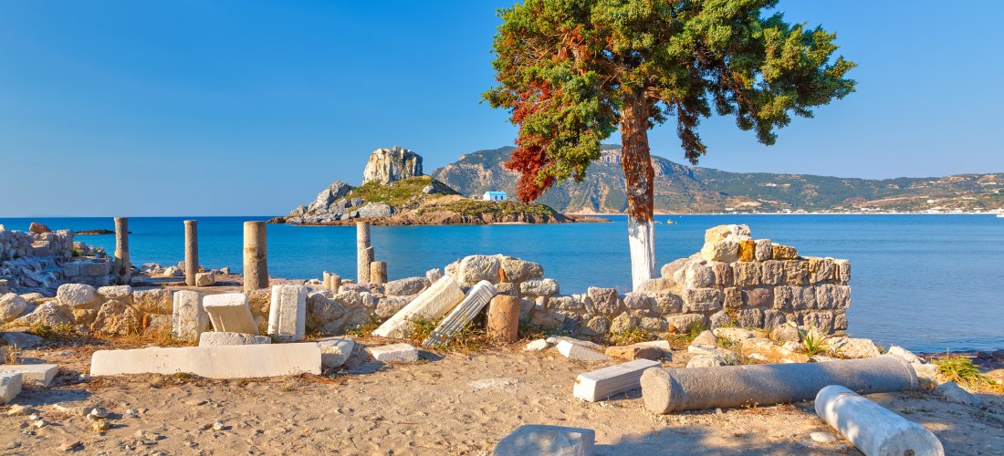 Antike Ruinen mit verstreut liegenden Säulentrommeln und Mauerresten am Meer. Im Vordergrund steht ein einzelner Baum mit grün-rotem Laub. Dahinter liegt eine kleine Insel mit einer weißen Kapelle auf einem Felsen, umgeben von tiefblauem Wasser. Im Hinter
