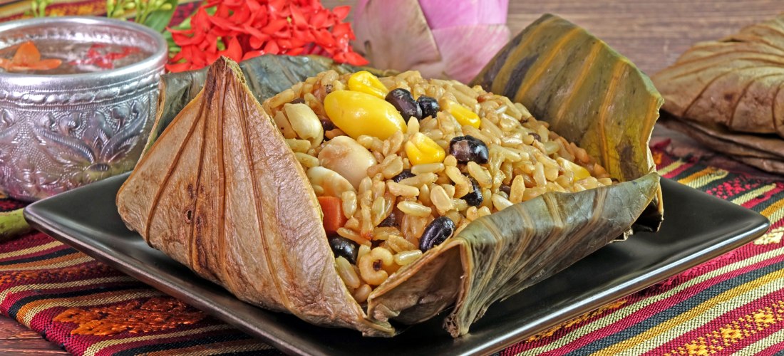 Traditionelles thailändisches Gericht aus gekochtem Reis mit Bohnen, Mais und Nüssen, serviert in einem gefalteten Lotusblatt auf einem schwarzen Teller. Im Hintergrund thailändische Stoffdeko, Blüten und eine verzierte Silberschale.