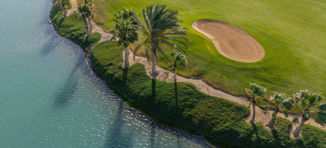 Luftaufnahme eines gepflegten Golfplatzes mit grüner Rasenfläche und Palmen, die sich entlang eines ruhigen Gewässers erstrecken. Rechts ist ein Sandbunker sichtbar, links das türkisfarbene Wasser mit leichtem Wellenschlag.