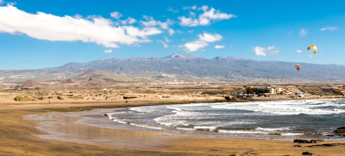 langer Naturstrand mit goldbraunem Sand, Wellen, Dünenlandschaft und Kitesurfern am Himmel.