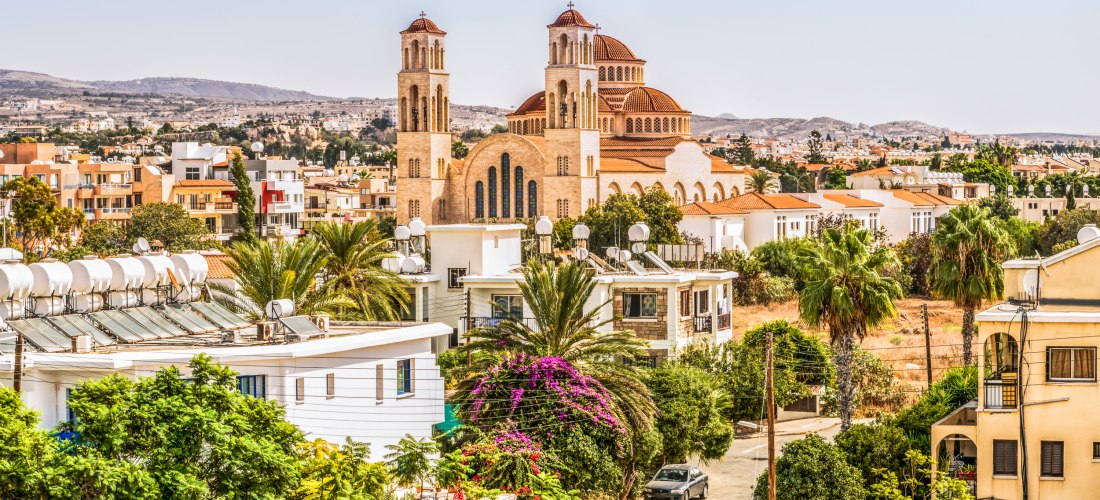 Blick auf die Agioi Anargyroi Kirche in Paphos mit rotbraunen Kuppeln, Palmen und Wohnhäusern im Vordergrund.