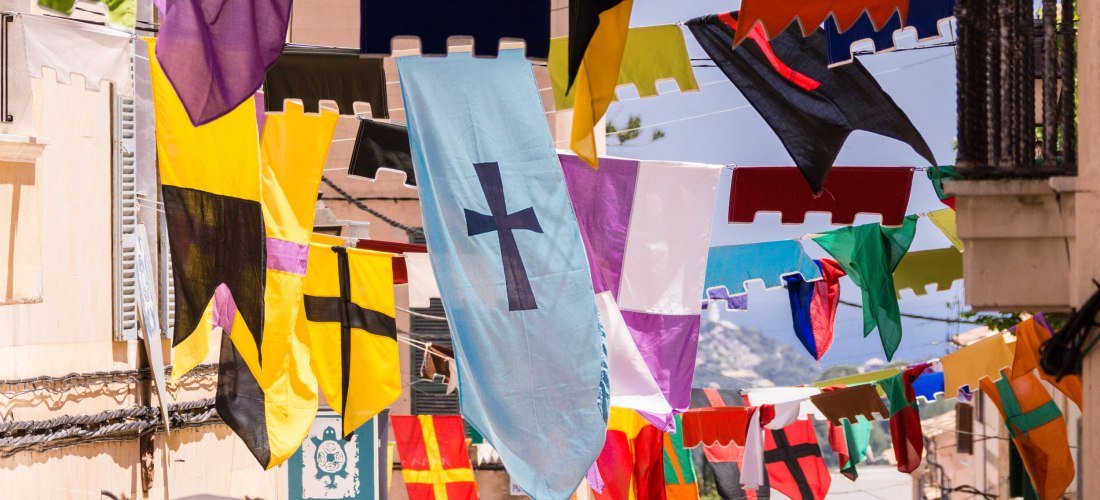 Bunte Fahnen und Wimpel hängen quer über einer Altstadtstraße. Sie tragen Kreuze und einfache Muster in leuchtenden Farben wie Gelb, Rot, Blau, Schwarz und Lila. Im Hintergrund sind Häuserfassaden, Fensterläden und ein Firmenschild mit der Aufschrift 'Tor