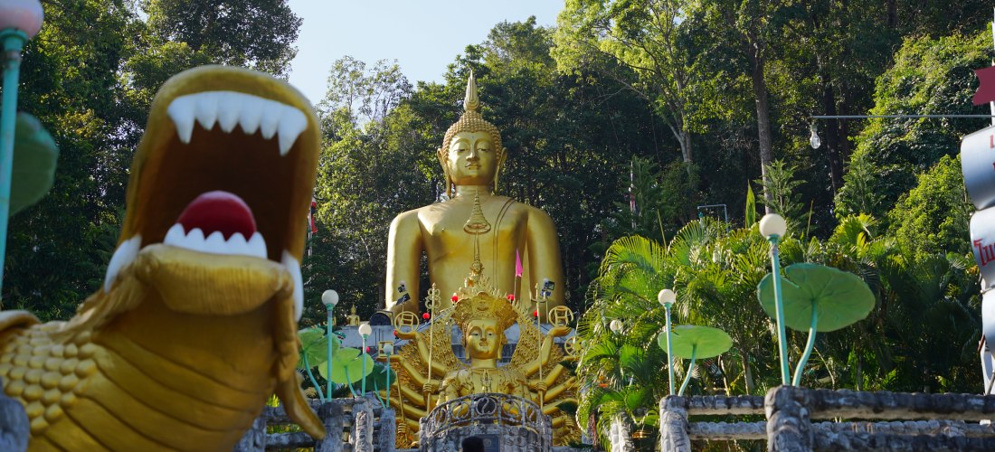 Das Bild zeigt eine imposante goldene Buddha-Statue, die auf einem von Palmen und Dschungelvegetation umgebenen Hügel thront. Im Vordergrund führt eine breite Treppe mit Geländern aus Stein zu der Statue hinauf. Links ist eine große, goldene Drachenfigur 
