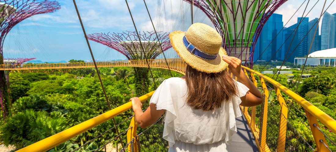 Eine Frau in weißem Sommerkleid und Strohhut steht auf einem erhöhten, gelb-geländerten Skywalk im Gardens by the Bay in Singapur. Sie blickt auf die ikonischen Supertrees – riesige, futuristische Baumskulpturen mit vertikalen Gärten und filigranen Metall